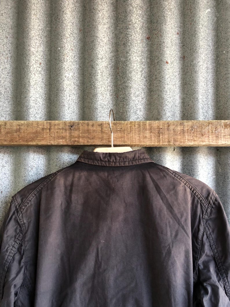 May include: A dark brown jacket hanging on a wooden bar. The jacket has a collar and visible stitching along the seams. The background is a corrugated metal sheet.