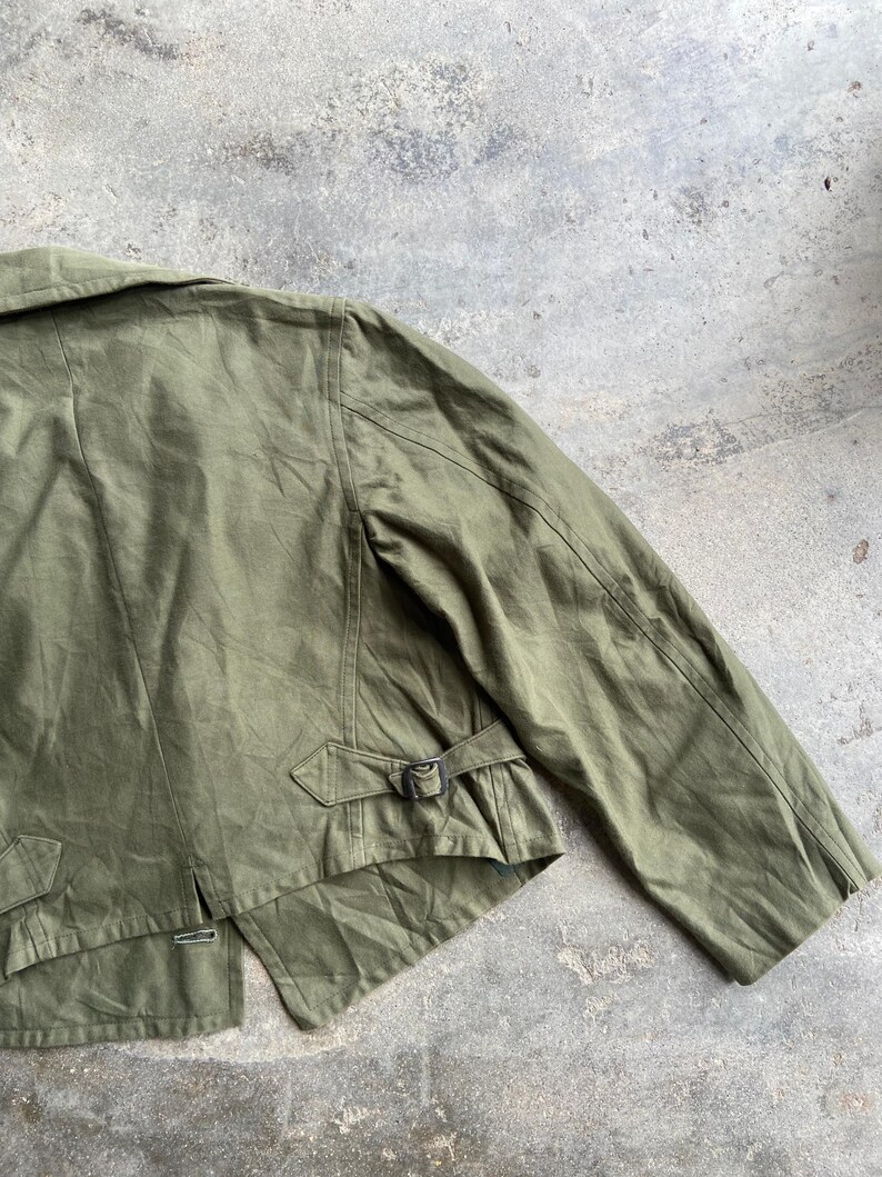 May include: Olive green jacket with a notched collar, shoulder seams, and a belt detail at the waist. The jacket has a classic design with a button closure and a tailored fit. The fabric appears to be a durable cotton or blend, suitable for various weather conditions.