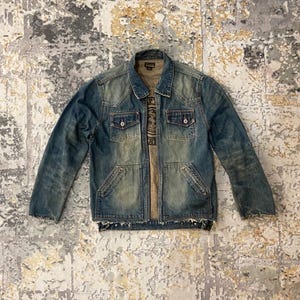 May include: A faded blue denim jacket with a collared neckline, two chest pockets, and two lower pockets. The jacket has a distressed look with frayed edges and a button-up front. A graphic tee is visible underneath.