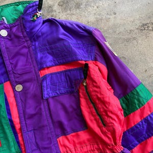 May include: A vintage ski jacket in purple, red, green, and black. The jacket features a high collar, multiple pockets, and a zip closure. It has a retro colour block design with the brand logo on the sleeve.