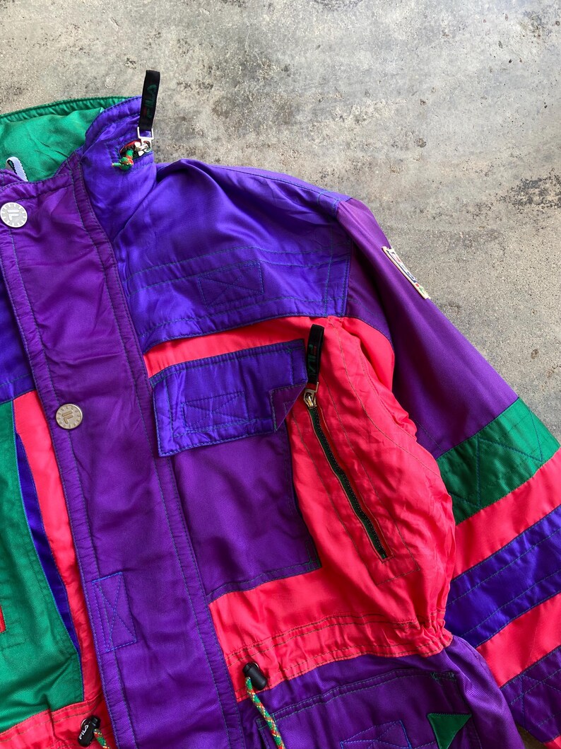 May include: A vintage ski jacket in purple, red, green, and black. The jacket features a high collar, multiple pockets, and a zip closure. It has a retro colour block design with the brand logo on the sleeve.
