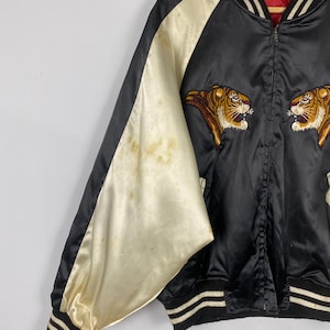 Puede incluir: Chaqueta bomber de sat&eacute;n negro con mangas color crema y cabezas de tigre bordadas. La chaqueta tiene cuello, pu&ntilde;os y dobladillo de canal&eacute; con rayas blancas. Forro rojo y cierre de cremallera.