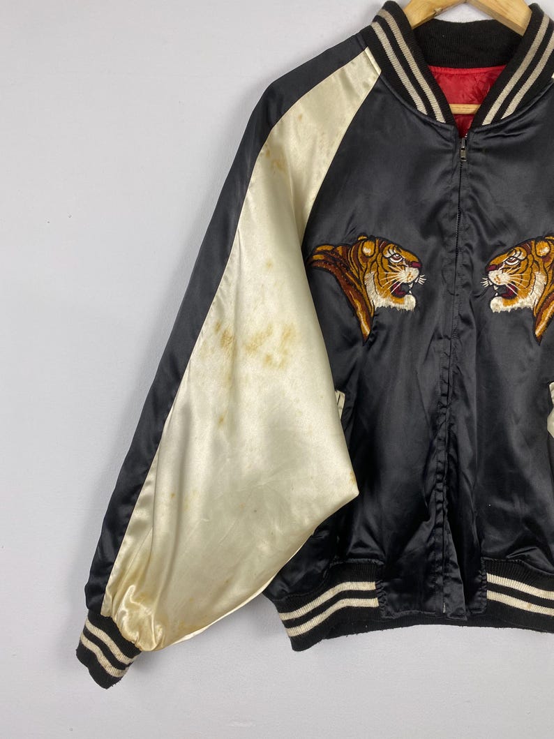 Puede incluir: Chaqueta bomber de sat&eacute;n negro con mangas color crema y cabezas de tigre bordadas. La chaqueta tiene cuello, pu&ntilde;os y dobladillo de canal&eacute; con rayas blancas. Forro rojo y cierre de cremallera.