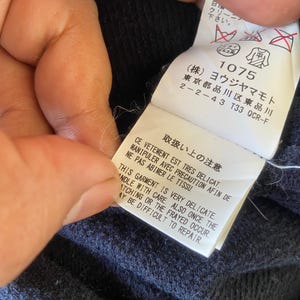 Pu&ograve; includere: Primo piano di un'etichetta di abbigliamento blu navy con testo giapponese e simboli di cura. L'etichetta include il testo "THIS GARMENT IS VERY DELICATE. HANDLE WITH CARE. ALSO ONCE THE SNATCHING OR THE FRAYED OCCUR. MAY BE DIFFICULT TO REPAIR."