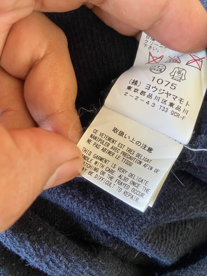 Pu&ograve; includere: Primo piano di un'etichetta di abbigliamento blu navy con testo giapponese e simboli di cura. L'etichetta include il testo "THIS GARMENT IS VERY DELICATE. HANDLE WITH CARE. ALSO ONCE THE SNATCHING OR THE FRAYED OCCUR. MAY BE DIFFICULT TO REPAIR."