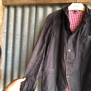 May include: A dark brown blazer with a red and white checkered lining. The blazer has three buttons, two front pockets, and a notched lapel. The jacket is hanging on a hanger with the brand name "MANGO" on it.