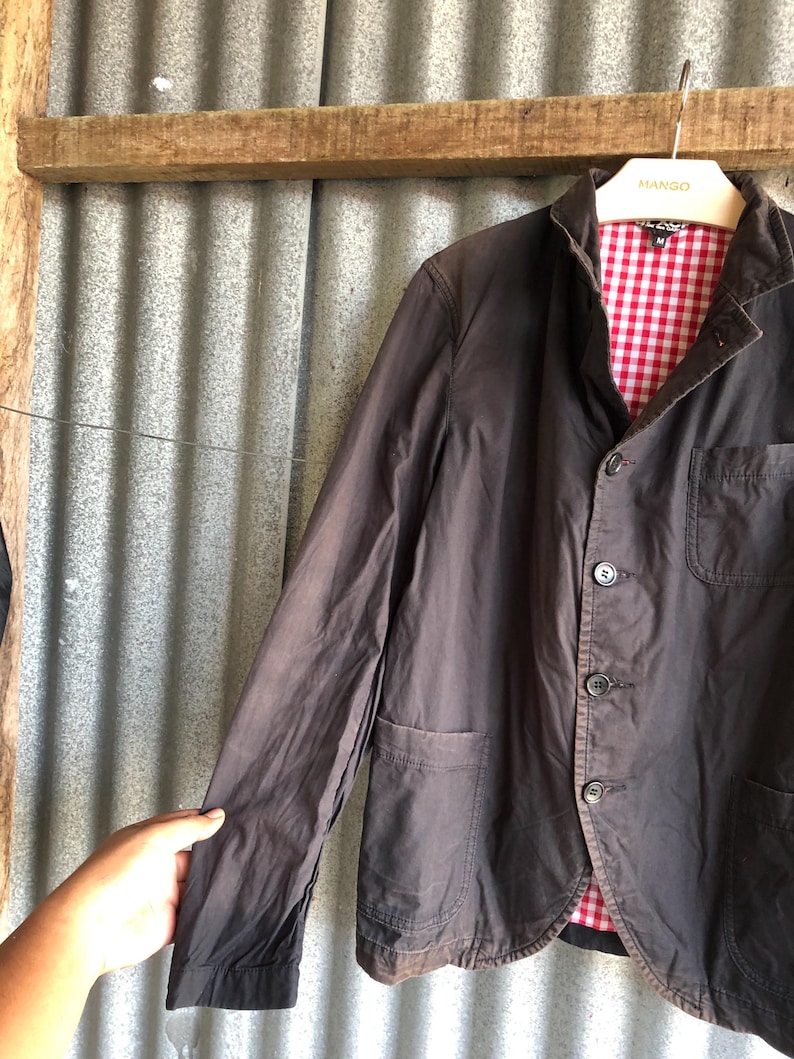 May include: A dark brown blazer with a red and white checkered lining. The blazer has three buttons, two front pockets, and a notched lapel. The jacket is hanging on a hanger with the brand name "MANGO" on it.