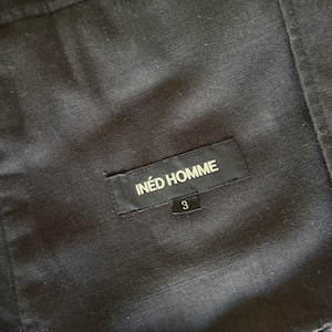 May include: A black fabric garment featuring a rectangular black label stitched onto the material. The label displays the white text IN&Eacute;D HOMME with a smaller black tag below it showing the white number 3. The fabric has a visible woven texture.
