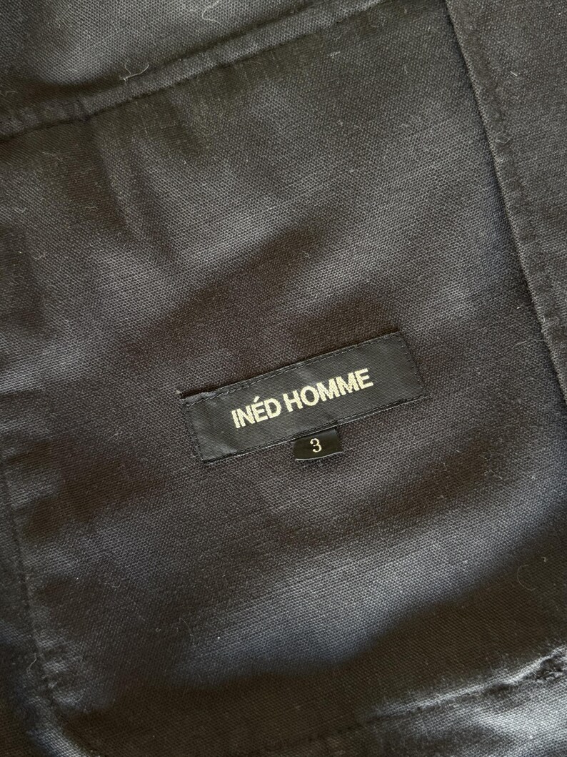 May include: A black fabric garment featuring a rectangular black label stitched onto the material. The label displays the white text IN&Eacute;D HOMME with a smaller black tag below it showing the white number 3. The fabric has a visible woven texture.