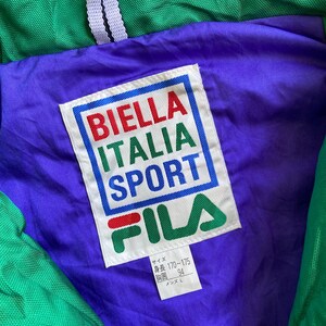 May include: Close-up of a vintage green, purple, and red Fila jacket. The jacket's interior features a purple lining with a white label displaying "BIELLA ITALIA SPORT" in red, green, and blue. The Fila logo is visible below the text.