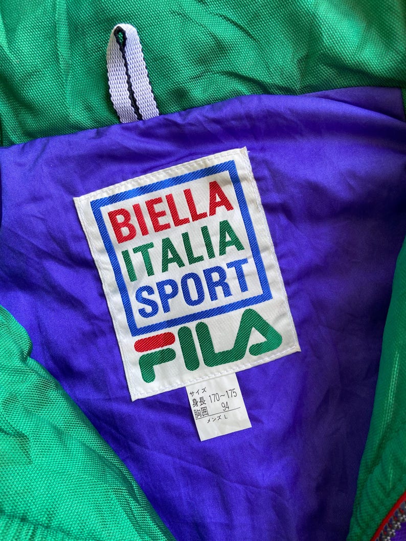 May include: Close-up of a vintage green, purple, and red Fila jacket. The jacket's interior features a purple lining with a white label displaying "BIELLA ITALIA SPORT" in red, green, and blue. The Fila logo is visible below the text.