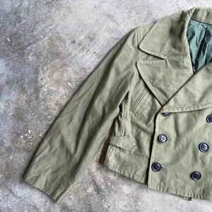 May include: Olive green double-breasted jacket with black buttons. The jacket features a notched collar, two front pockets, and a tailored fit. The sleeve is visible on the left side. The jacket is made of a durable fabric.