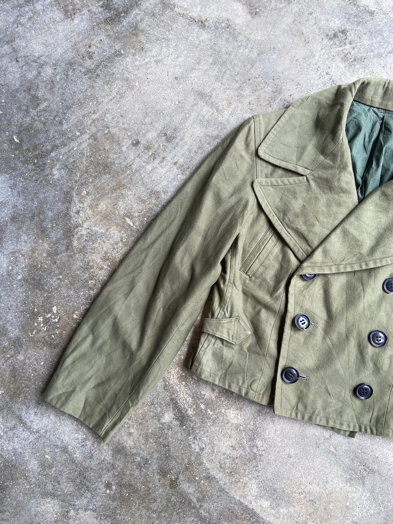 May include: Olive green double-breasted jacket with black buttons. The jacket features a notched collar, two front pockets, and a tailored fit. The sleeve is visible on the left side. The jacket is made of a durable fabric.