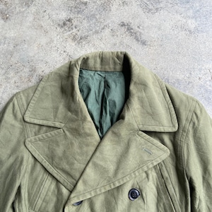 May include: Olive green double-breasted coat with a notched collar and four dark buttons. The coat has a tailored fit and a green lining visible at the neckline. The fabric appears to be a durable, possibly military-style material.