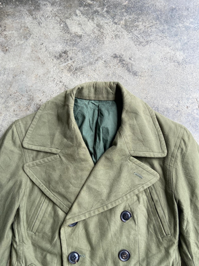 May include: Olive green double-breasted coat with a notched collar and four dark buttons. The coat has a tailored fit and a green lining visible at the neckline. The fabric appears to be a durable, possibly military-style material.