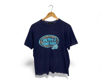 May include: Navy blue t-shirt with a graphic design featuring the text "Long Wave Sandy Beach" in a stylized oval shape. The design includes blue and turquoise accents. The t-shirt is hanging on a wooden hanger.