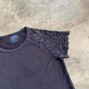 Pu&ograve; includere: T-shirt nero con girocollo e dettaglio in pizzo sulla spalla destra. La maglietta ha un'etichetta con la scritta "REGULATION" e una firma. La maglietta &egrave; realizzata in materiale morbido.