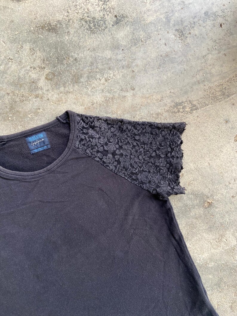 Pu&ograve; includere: T-shirt nero con girocollo e dettaglio in pizzo sulla spalla destra. La maglietta ha un'etichetta con la scritta "REGULATION" e una firma. La maglietta &egrave; realizzata in materiale morbido.