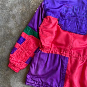 May include: A vintage colour-blocked jacket in shades of purple, pink, and green. The jacket features the brand name "FILA" in purple on the upper back. The jacket has a zip-up front and a high collar. The sleeves have a ribbed cuff.
