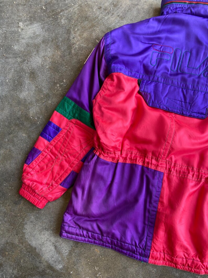 May include: A vintage colour-blocked jacket in shades of purple, pink, and green. The jacket features the brand name "FILA" in purple on the upper back. The jacket has a zip-up front and a high collar. The sleeves have a ribbed cuff.