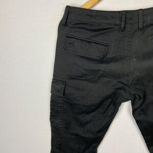 May include: Black cargo trousers with two side pockets and a back pocket, hanging on a wooden hanger. The trousers are made of a solid, dark fabric.