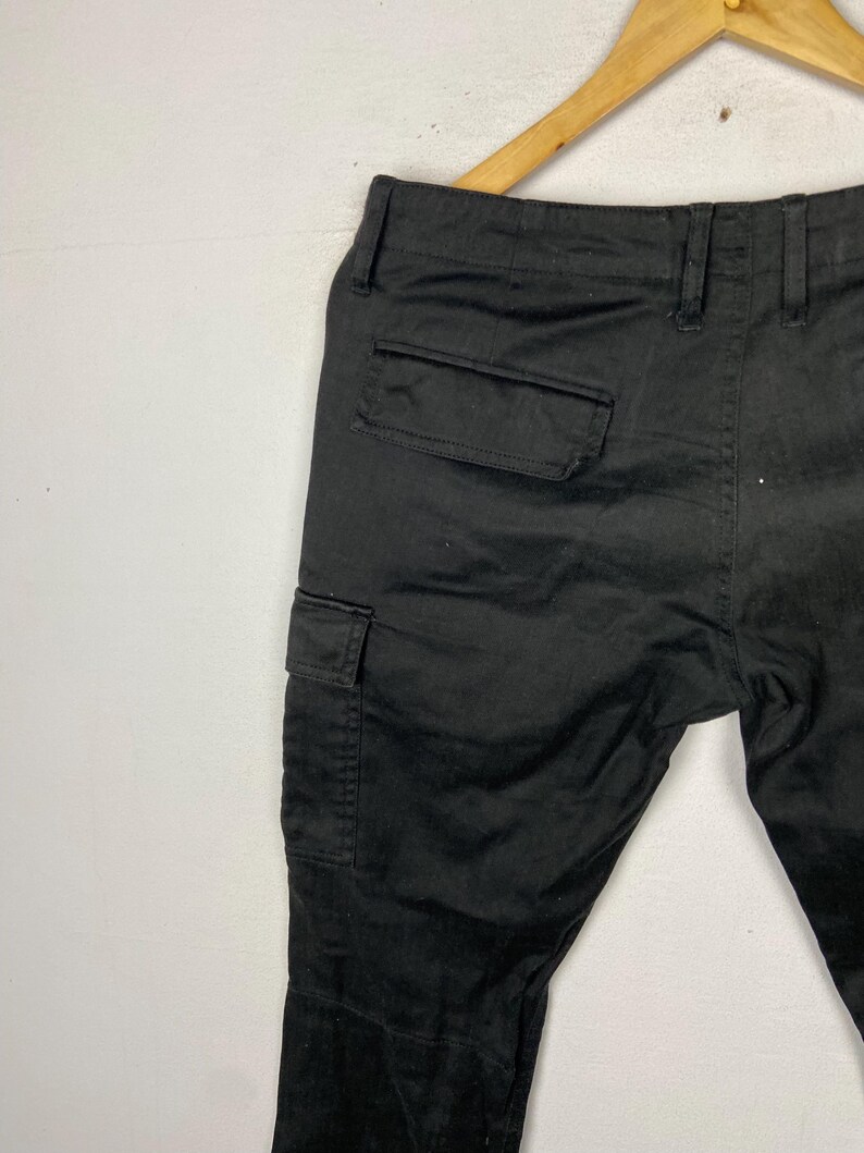 May include: Black cargo trousers with two side pockets and a back pocket, hanging on a wooden hanger. The trousers are made of a solid, dark fabric.