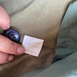 May include: Close-up of a dark blue button on olive green fabric. A small white tag with Japanese text is visible. The fabric has a subtle texture and a slight sheen. The button is round with four holes.