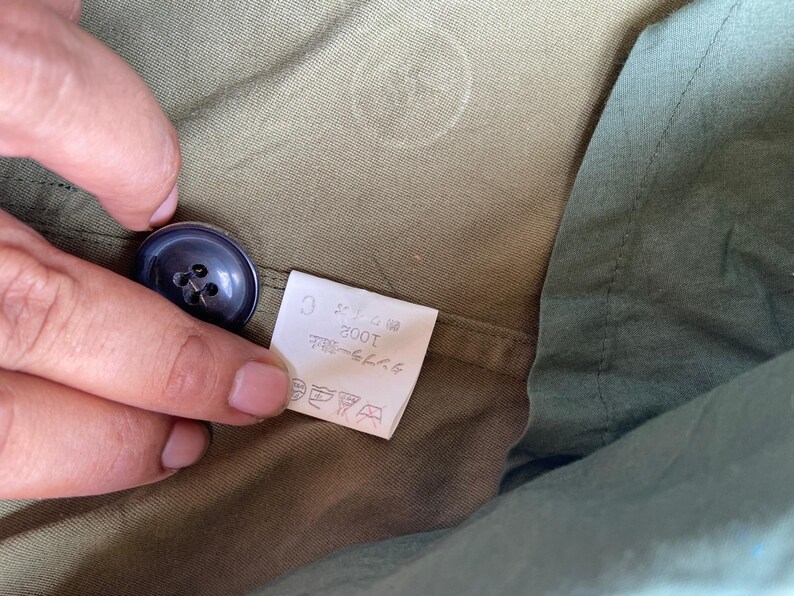 May include: Close-up of a dark blue button on olive green fabric. A small white tag with Japanese text is visible. The fabric has a subtle texture and a slight sheen. The button is round with four holes.