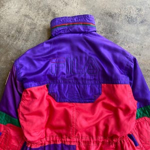 May include: A vintage ski suit in purple, red, and green. The suit features the brand name "FILA" embroidered on the back. The collar has a striped design with green, red, and yellow. The suit is made of a shiny material, possibly nylon.