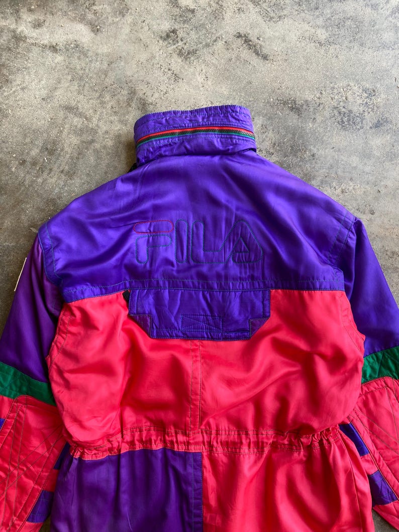 May include: A vintage ski suit in purple, red, and green. The suit features the brand name "FILA" embroidered on the back. The collar has a striped design with green, red, and yellow. The suit is made of a shiny material, possibly nylon.