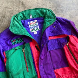 May include: A vintage Fila Italia Sport jacket in purple, green, and red. The jacket features a high collar, multiple pockets with zippers, and the Fila logo. The jacket has a drawstring at the waist and the text "BIELLA ITALIA SPORT" on the label.