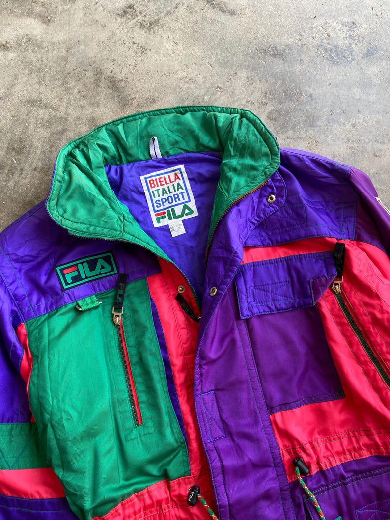 May include: A vintage Fila Italia Sport jacket in purple, green, and red. The jacket features a high collar, multiple pockets with zippers, and the Fila logo. The jacket has a drawstring at the waist and the text "BIELLA ITALIA SPORT" on the label.