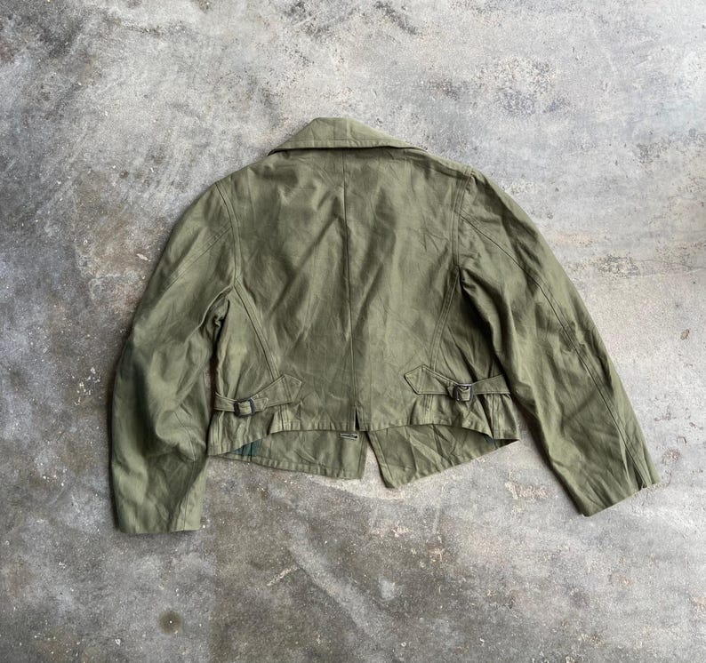 May include: Olive green jacket with a collar and two small buckles on the back. The jacket has a tailored fit and a classic design. The fabric appears to be a lightweight material, suitable for layering.