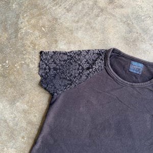 Pu&ograve; includere: Una maglietta grigio scuro con girocollo e manica in pizzo. La manica in pizzo &egrave; di una tonalit&agrave; di grigio pi&ugrave; scura e presenta un motivo floreale. La maglietta ha un'etichetta sul retro.