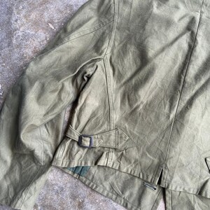 May include: Olive green jacket with a collar and a belt detail at the waist. The jacket has a tailored fit and a button closure. The fabric appears to be a durable material, suitable for outerwear. The jacket's design is classic and functional.