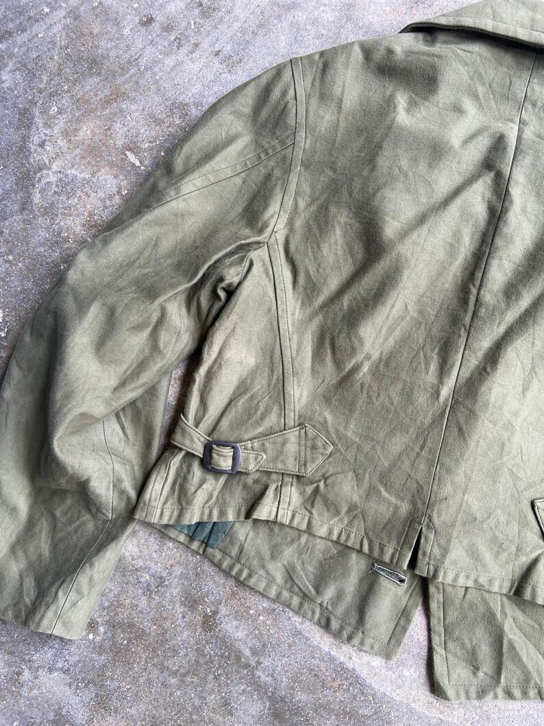 May include: Olive green jacket with a collar and a belt detail at the waist. The jacket has a tailored fit and a button closure. The fabric appears to be a durable material, suitable for outerwear. The jacket's design is classic and functional.