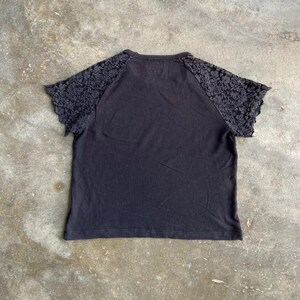Pu&ograve; includere: T-shirt nero con maniche in pizzo. La maglietta a maniche corte ha uno scollo tondo e un corpo nero a tinta unita. Le maniche sono in pizzo grigio scuro. La maglietta &egrave; stesa su una superficie di cemento.