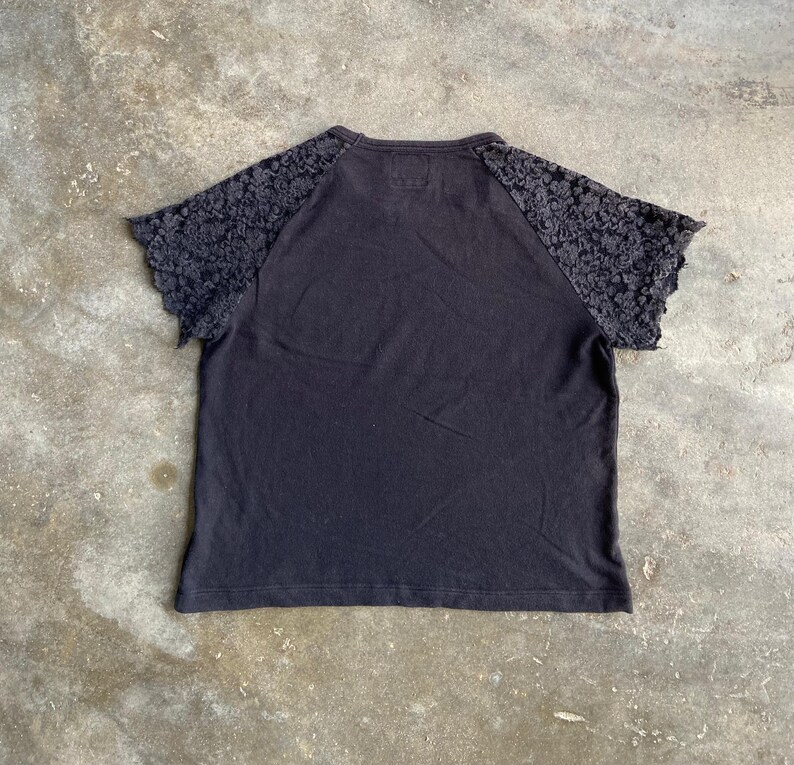 Pu&ograve; includere: T-shirt nero con maniche in pizzo. La maglietta a maniche corte ha uno scollo tondo e un corpo nero a tinta unita. Le maniche sono in pizzo grigio scuro. La maglietta &egrave; stesa su una superficie di cemento.
