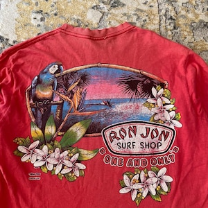 May include: Red t-shirt featuring a tropical scene with a parrot, flowers, and a sunset over the ocean. The shirt has the text "Ron Jon Surf Shop One and Only" in a circular logo design.