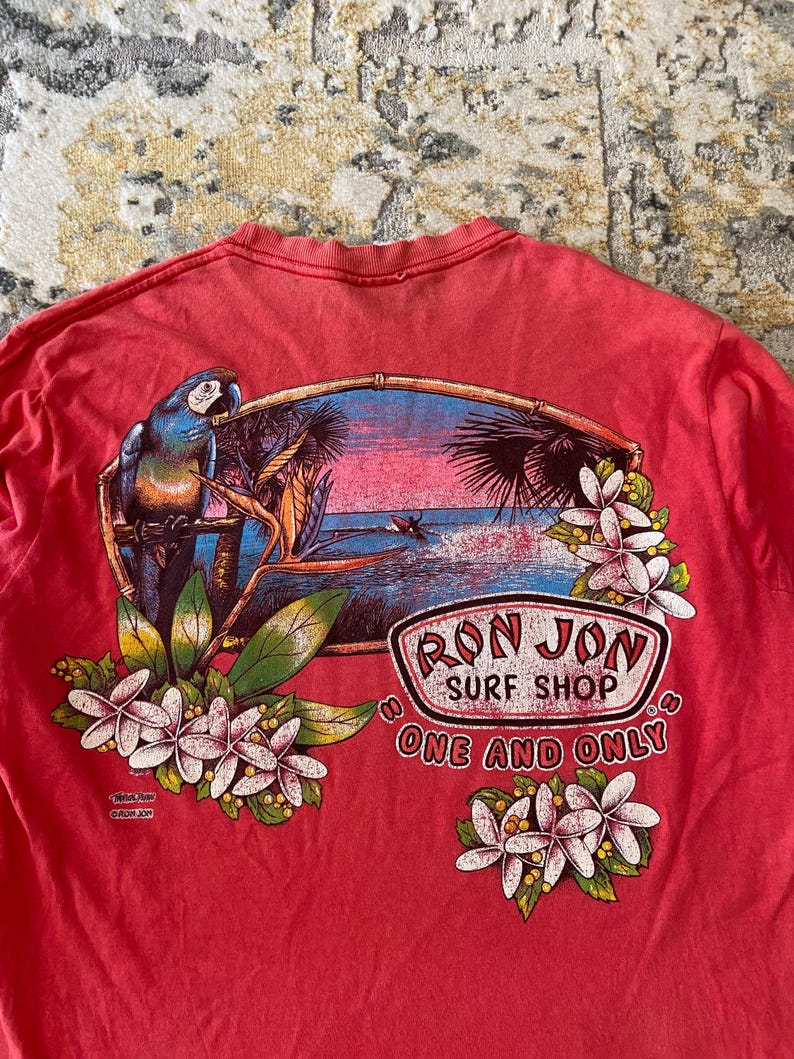 May include: Red t-shirt featuring a tropical scene with a parrot, flowers, and a sunset over the ocean. The shirt has the text "Ron Jon Surf Shop One and Only" in a circular logo design.