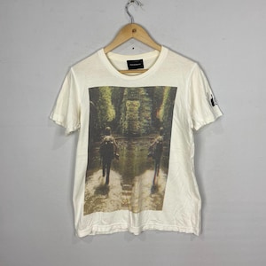 May include: White t-shirt with a graphic print of two people walking through a forest. The print is in shades of green, brown, and blue. The t-shirt has a crew neck and short sleeves.