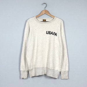 Może przedstawiać: Jasnobeżowa bluza z długimi rękawami i okrągłym dekoltem. Bluza ma czarny nadruk graficzny "USAF" z przodu.