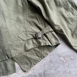 May include: Close-up of an olive green jacket. The jacket features a buckle closure at the waist and detailed stitching. The fabric appears to be a durable, possibly canvas material. The jacket's design suggests a military or utilitarian aesthetic.