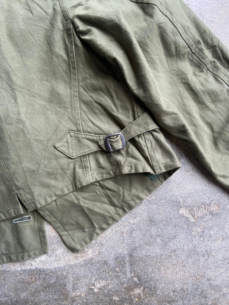 May include: Close-up of an olive green jacket. The jacket features a buckle closure at the waist and detailed stitching. The fabric appears to be a durable, possibly canvas material. The jacket's design suggests a military or utilitarian aesthetic.