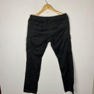 May include: Black cargo trousers hanging on a wooden hanger. The trousers have two pockets on the back and two cargo pockets on the sides. The trousers are made of a solid black material.