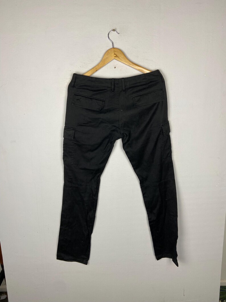 May include: Black cargo trousers hanging on a wooden hanger. The trousers have two pockets on the back and two cargo pockets on the sides. The trousers are made of a solid black material.