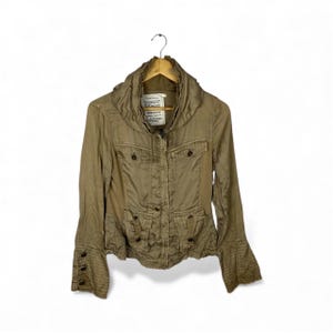 May include: A tan jacket with a high collar, multiple pockets, and button details. The jacket is hanging on a wooden hanger against a white background. The sleeves have a flared design.