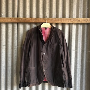 May include: A dark brown blazer with a red and white checkered lining, hanging on a metal hanger. The blazer has three buttons and two front pockets. The background is a corrugated metal wall.