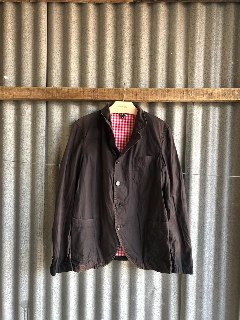 May include: A dark brown blazer with a red and white checkered lining, hanging on a metal hanger. The blazer has three buttons and two front pockets. The background is a corrugated metal wall.