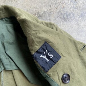 May include: Olive green jacket with a black label that reads "Y's" in white. The jacket has a button closure and a dark green lining. The fabric appears to be a durable, possibly cotton blend.